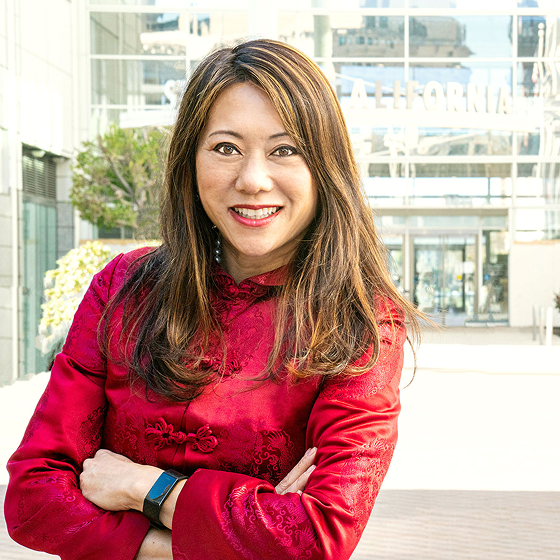 Fiona Ma, California State Treasurer, smiling and standing with her arms crossed, wearing a red traditional jacket with a subtle floral pattern, outdoors in front of a modern glass building.
