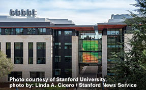 Exterior view of a modern Stanford University building with light stone cladding and dark trim, featuring a central glass section with a colorful, multicolored display visible inside. (Note: Text at the bottom provides photo credit.)